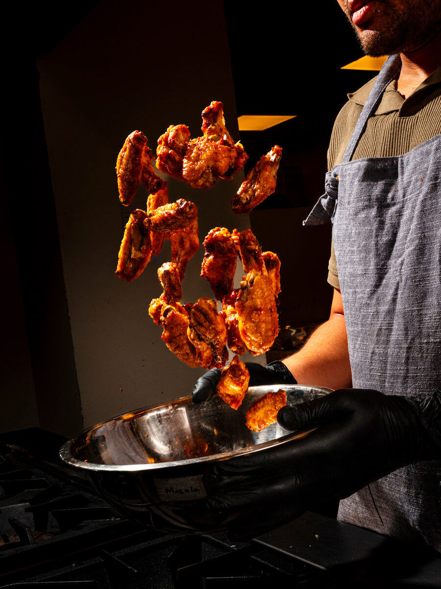 Masala wings mid-toss over a steel bowl in the kitchen
